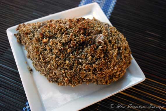 A single breast of Clean Eating Nut Crusted Baked Chicken sitting on a square, white plate and a dark brown, bamboo placemat.