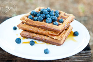 A white plate holds a stack of three homestyle waffles that are topped with fresh blueberries.