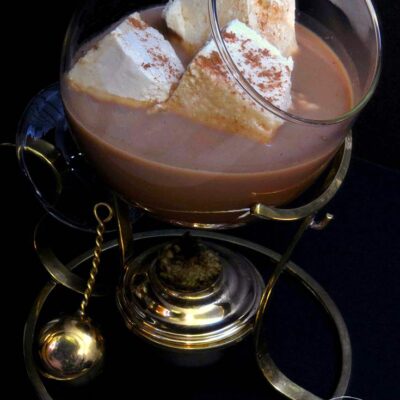 A brandy warmer sits filled with hot chocolate and three square marshmallows.