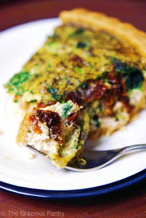 A slice of Clean Eating Pesto Quiche With Sun Dried Tomatoes sitting on a white plate. In front of the piece of quiche rests a fork with a piece of the quiche on it, ready to eat and enjoy.