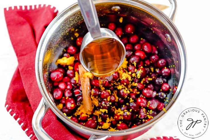 Pouring honey into a pot filled with fresh cranberries, orange zest and a cinnamon stick.