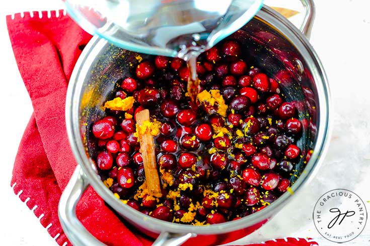 Pouring water into a pot filled with fresh cranberries, orange zest, honey and a cinnamon stick.