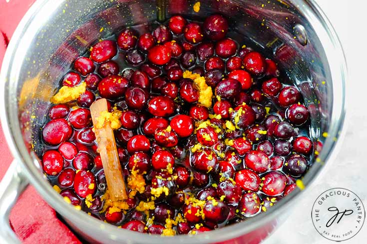 Simmering a pot filled with fresh cranberries, orange zest, honey, a cinnamon stick and water.