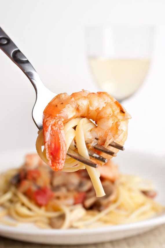 Clean Eating Shrimp Scampi In this image, the camera focuses on a fork being held with a small bit of pasts wrapped around it and a large shrimp. In the background, you see a bowl of Clean Eating Shrimp Scampi and a glass of white wine.