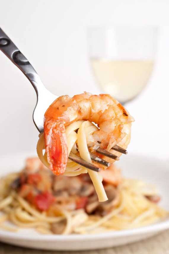 In this image, the camera focuses on a fork being held with a small bit of pasts wrapped around it and a large shrimp. In the background, you see a bowl of Clean Eating Shrimp Scampi and a glass of white wine.