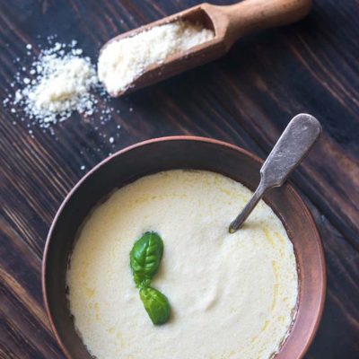 Healthy Alfredo Sauce in a serving bowl with a scoop of parmesan sitting to the side of the bowl.