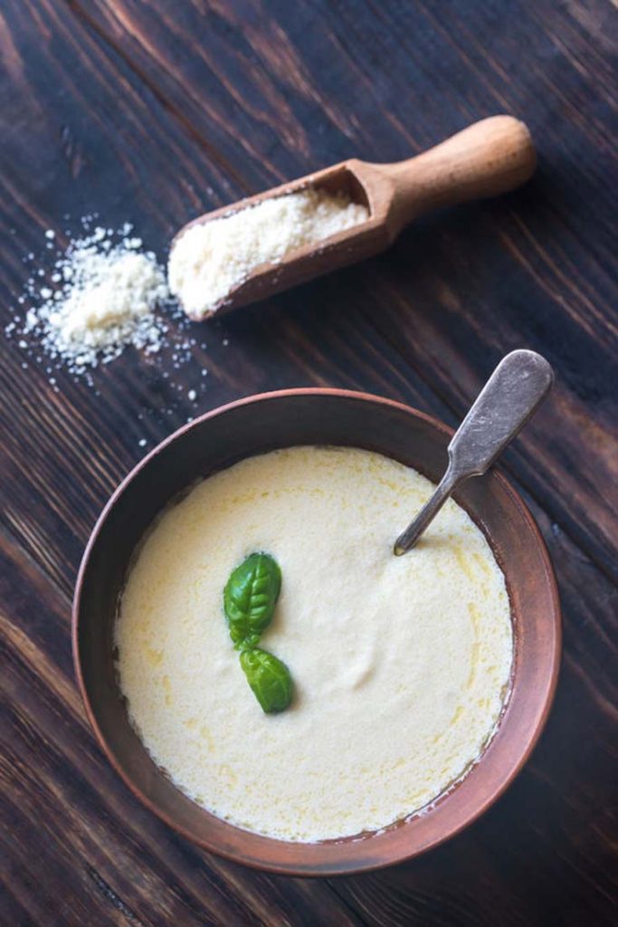 Healthy Alfredo Sauce in a serving bowl with a scoop of parmesan sitting to the side of the bowl.