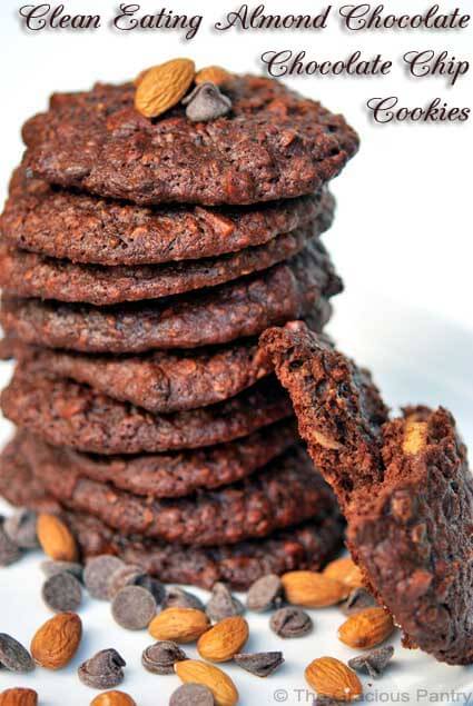 A larget stack of almond chocolate chocolate chip cookies sits on a white background. Chocolate chips and almonds are scattered around the base.