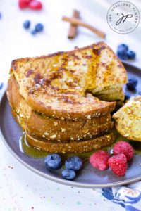 A stack of Healthy French Toast sits on a plate with fresh raspberries and blueberries. The toast is stacked and has a piece cut out of it.