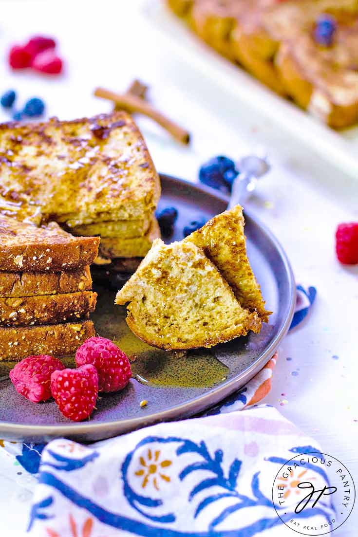 A slice of the stack of french toast sitting on a plate has been cut and rests on a fork on a plate next to the stack of french toast.