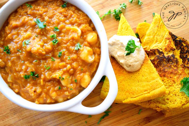 And overhead view of a bowl of this Clean Eating Garam Masala Red Lentil Stew. It's been sprinkled with a few freshly chopped herbs and has a side of flatbread wedges and hummus off to the right side of the bowl.