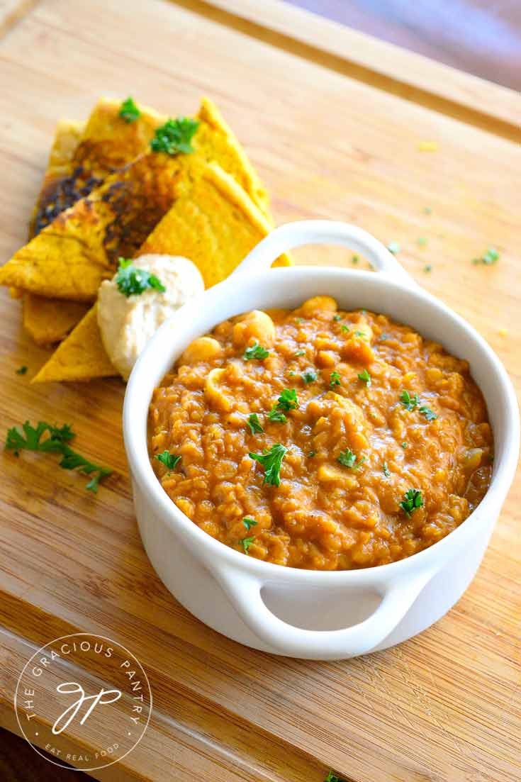 Clean Eating Garam Masala Red Lentil Stew sits in a white bowl on top of a large, wooden cutting board, served with flatbread wedges and a dollop of hummus.