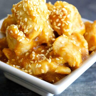 A pile of sesame chicken sits in a square, white bowl. You can see sesame seeds and delicious glaze coating the chicken in this sesame chicken recipe.