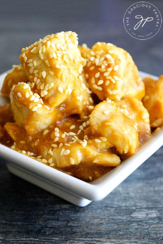 A pile of sesame chicken sits in a square, white bowl. You can see sesame seeds and delicious glaze coating the chicken in this sesame chicken recipe.
