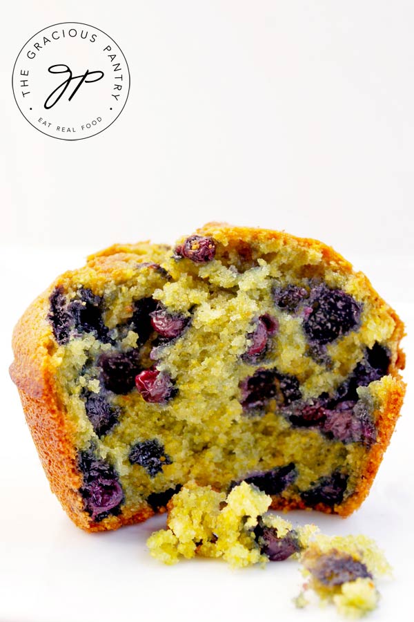 A single Blueberry Corn Muffin on a white background with the front half missing and a few crumbs laying in front of it.