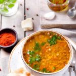 A bowl of this clean eating german lentil soup recipe sits on a white plate with some crackers on the side.