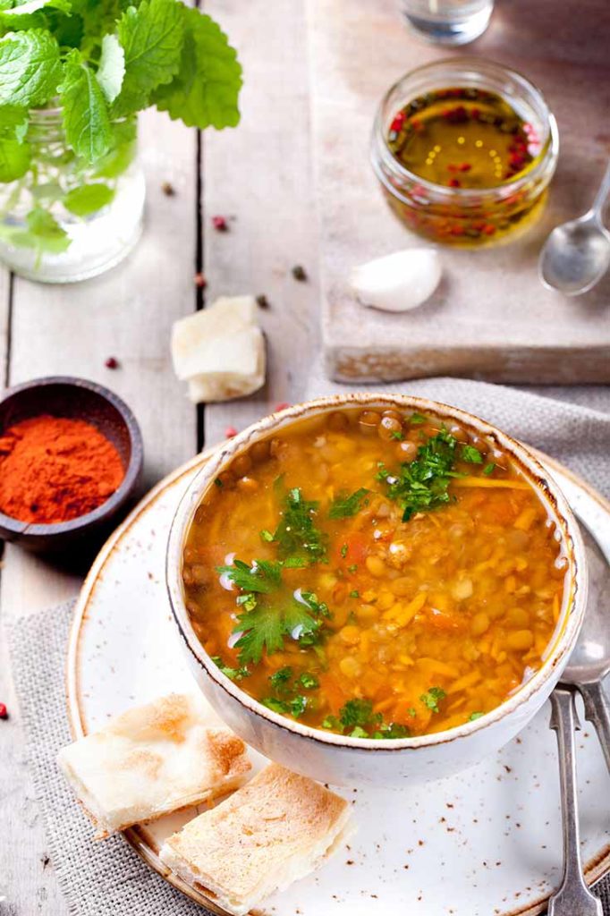 A bowl of this clean eating german lentil soup recipe sits on a white plate with some crackers on the side.