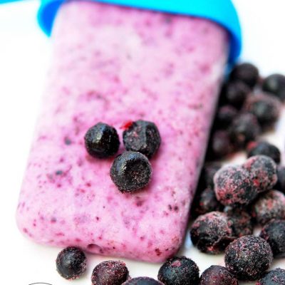 A single, Clean Eating Blueberry Yogurt Popsicles lays on a white background with frozen blueberries scattered all around it.