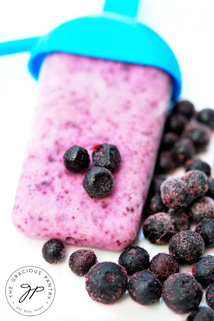 A single popsicle from this Clean Eating Blueberry Yogurt Popsicles recipe sits on a white background with blueberries sprinkled around it.