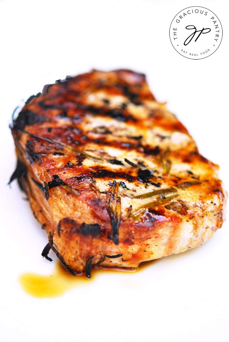 A single chops from this Clean Eating barbecue Rosemary Pork Chops recipe sits on a white background. It's beautifully caramelized with a tiny bit of the juices on the white plate.