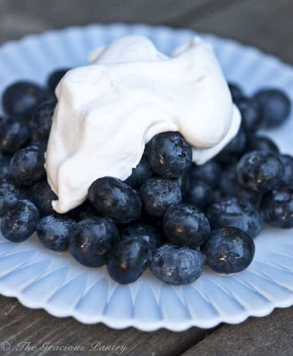 A plate of fresh blueberries is shown up close with this Clean Eating Coconut Whipped Cream laying over the top of them.