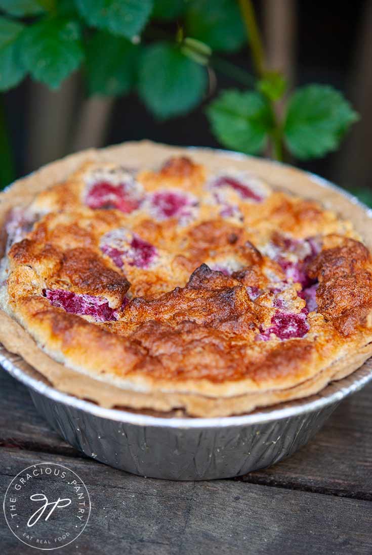 A just-baked Raspberry Pie sits on an outdoor bench, cooling. Green foliage sits behind it.