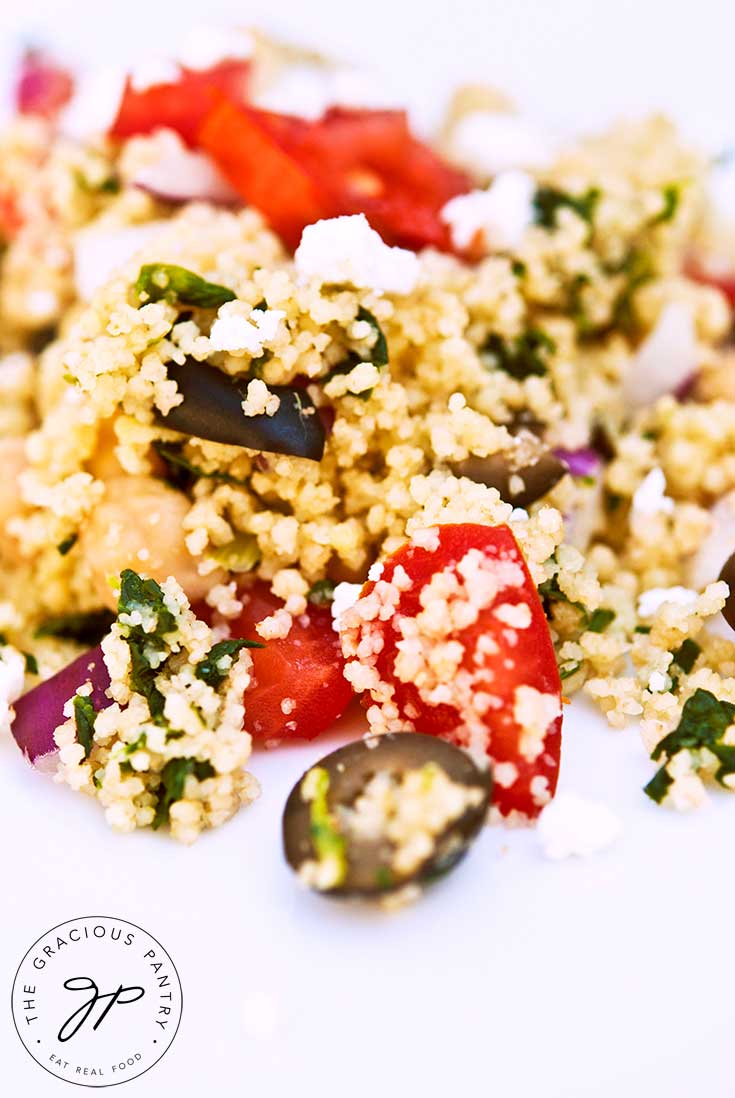 A large helping of this clean eating Greek couscous salad falls over the entirety of a white plate. You can see chunks of tomatoes, olives and feta cheese.