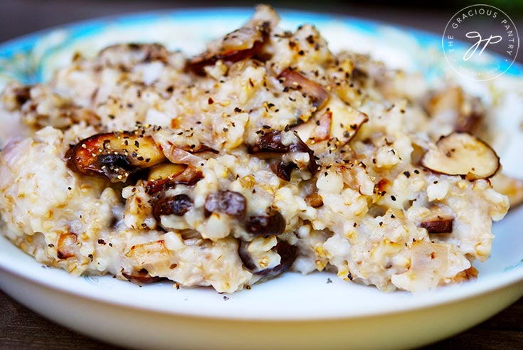 A bowl of this Clean Eating Savory Oatmeal sits on a dark background. You can see the golden color of the sautéed mushrooms  mixed into the oats.