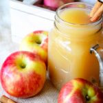 A clear, glass mug sits filled with this Apple Cider Recipe. It's surrounded by apples and cinnamon sticks. There are two cinnamon sticks in the jar for stirring the apple cider.