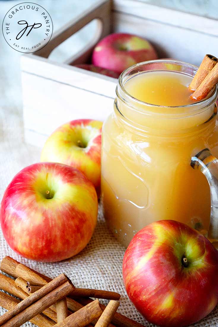 A clear, glass mug sits filled with this Apple Cider Recipe. It's surrounded by apples and cinnamon sticks. There are two cinnamon sticks in the jar for stirring the apple cider.