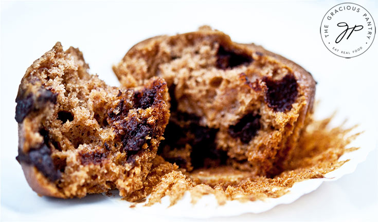 One of these Clean Eating Chocolate Chip Muffins is broken open to show off the lovely, chocolate chip filled center.
