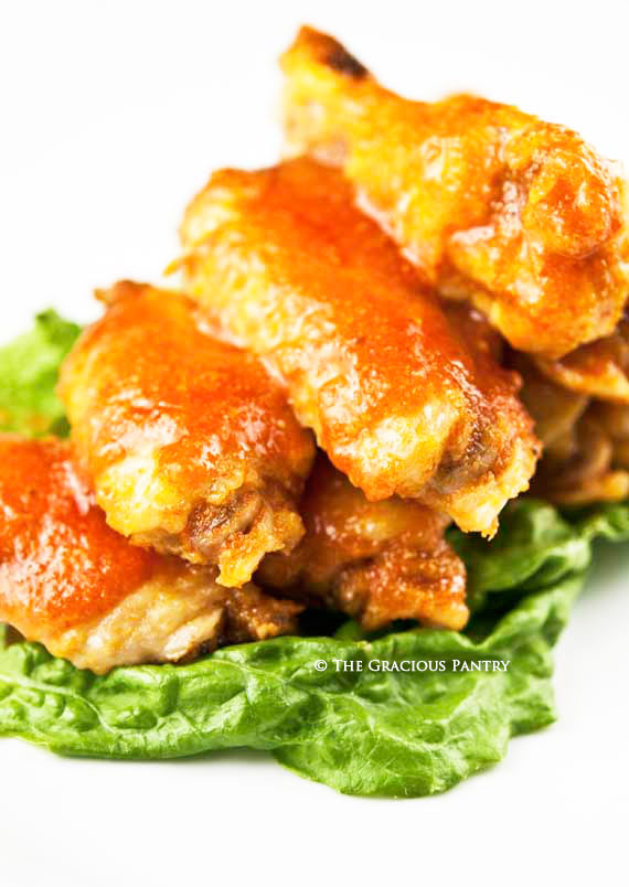 Clean Eating Spicy Buffalo Wings piled high on a bed of green lettuce and on a white serving platter. the red-orange buffalo sauce is nicely roasted onto the wings.