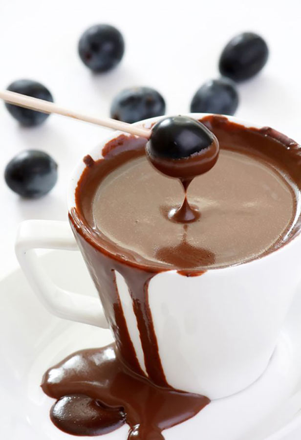 A single blueberry on a toothpick is being dipped into a white cup filled with Dark Chocolate Fondue.