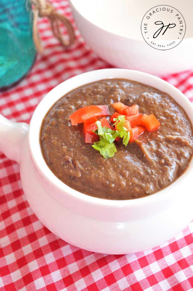 A white bowl of this Clean Eating Black Bean Soup sits on a red and white checkered table cloth and is garnished with bits of fresh, chopped tomato and a small sprig of fresh cilantro.