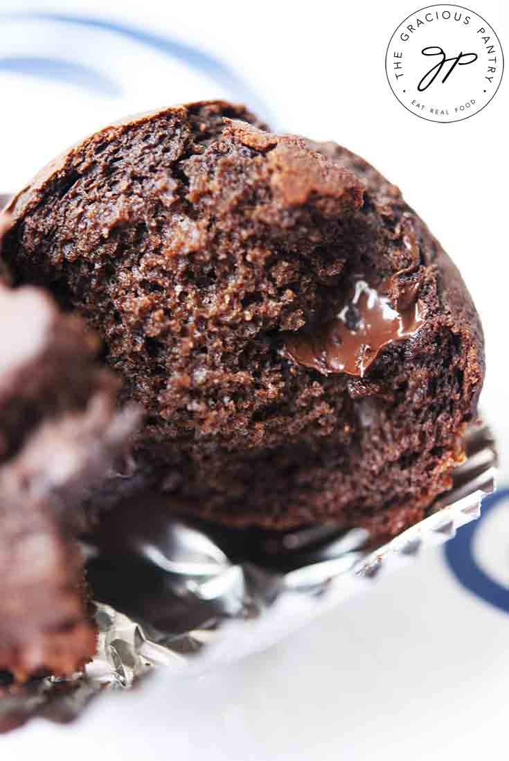 One of these Chocolate Chocolate Chip Muffins sits broken open, showing melty chocolate chips inside, warm out of the oven.