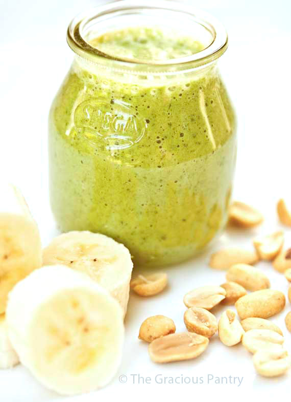 A small, clear, bless jar filled with this green, Clean Eating Peanut Butter And Banana Smoothie sits on a white background amidst sliced bananas and peanuts.