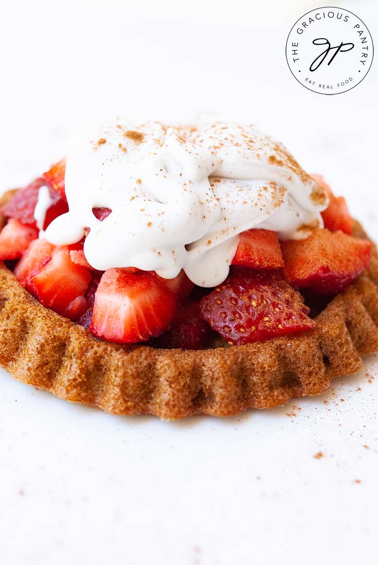 A strawberry shortcake sits on an all white background. There is a golden brown, cake base, a tall pile of strawberries and a good sized dollop of whopped cream. Cinnamon has been sprinkled over the top.