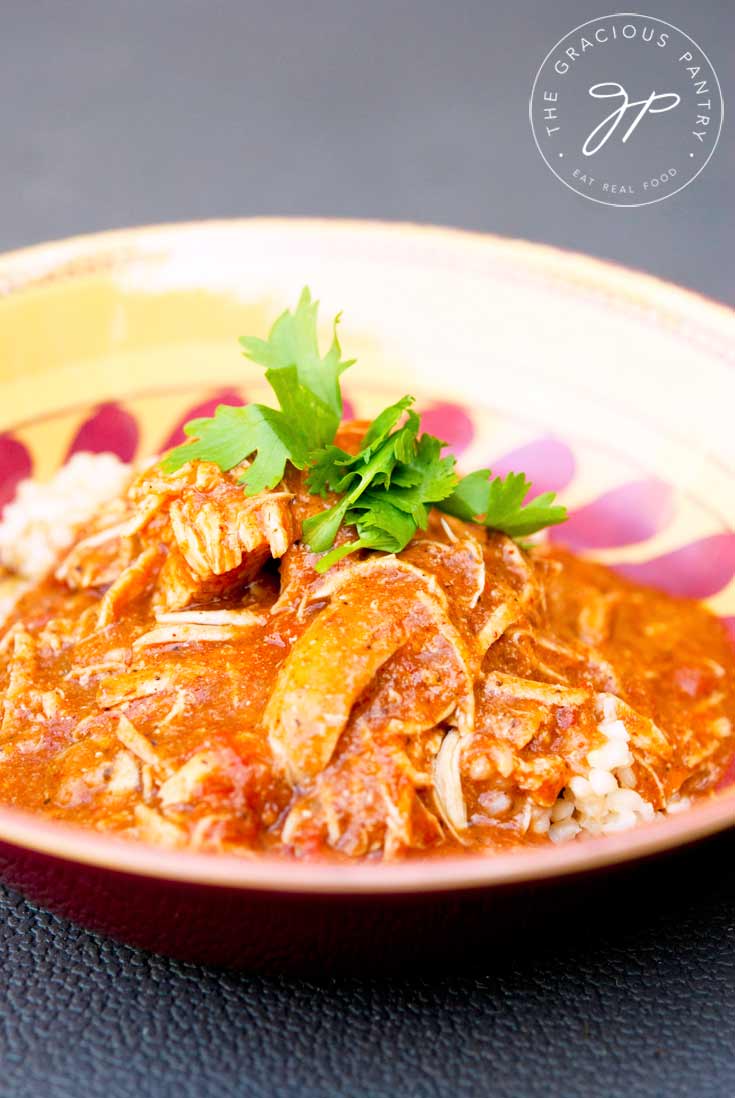 A bowl of Clean Eating Chicken Masala on a gray background. The red sauce with chicken sits ladled over brown rice.