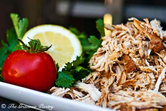 Shredded, Clean Eating Crock Pot Mexican Chicken served in a heap on a white platter. There is a whole tomato, bunch of cilantro and half a lemon on the platter for garnish.