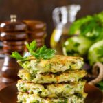 A stack of zucchini fritters sit on a wooden plate. There is an oil bottle and salt and pepper shaker sitting behind the plate. Fresh herbs top the fritters.
