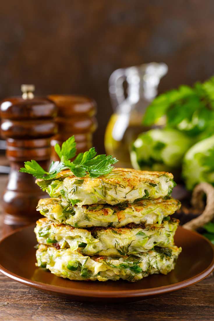 A stack of zucchini fritters sit on a wooden plate. There is an oil bottle and salt and pepper shaker sitting behind the plate. Fresh herbs top the fritters.