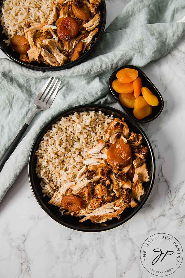 An overhead view of this Clean Eating Curry Chicken dish shows tow bowls filled with the curry chicken and brown rice. A small bowl of dried apricots sits to the side of these bowl on the right, with a fork on the left.