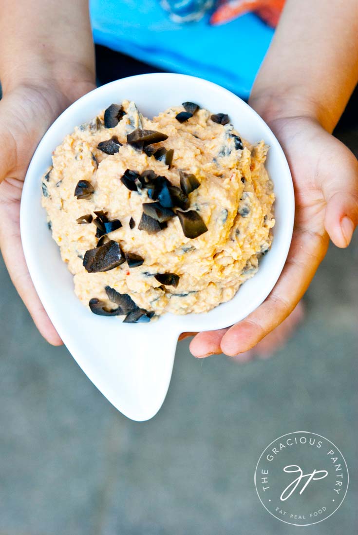 Pizza Hummus being held in a white bowl. It is topped with chopped, black olives.