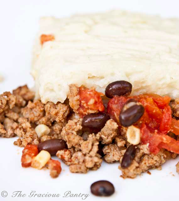 A servings of this Clean Eating Mexican Shepherds Pie sits on a rounds white plate on a white background. You can see the layers of the casserole with the white potato layer on top and the ground meat with beans on the bottoms.