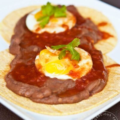 A plate of huevos rancheros sits on a table, ready to enjoy. It's layered with the tortillas first, then the beans and then the eggs on top with a bit of green garnish.