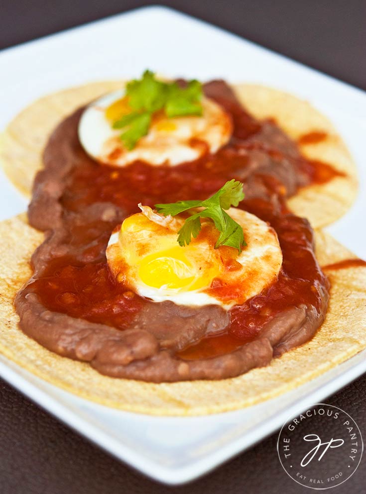 A plate of huevos rancheros sits on a table, ready to enjoy. It's layered with the tortillas first, then the beans and then the eggs on top with a bit of green garnish.