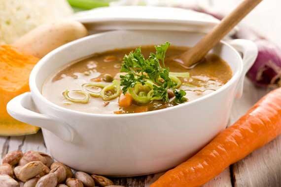 A bowl of Clean Eating "Build It Yourself" Vegetable Soup sits amidst fresh vegetables such as carrots and cut squash, ready to eat.