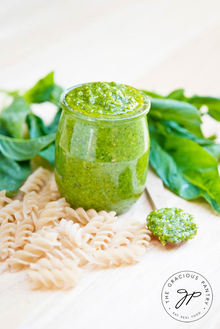 A small, clear jar of this Clean Eating Traditional Pesto Sauce sits on a light wood surface surrounded by dry, spiral pasta noodles and fresh basil leaves.