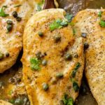 And up close view of this lemon caper chicken still sitting in the skillet with a serving spoon lifting up one of the chicken breasts. It has capers sprinkled over it as well as fresh parsley.