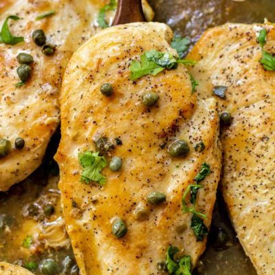 And up close view of this lemon caper chicken still sitting in the skillet with a serving spoon lifting up one of the chicken breasts. It has capers sprinkled over it as well as fresh parsley.